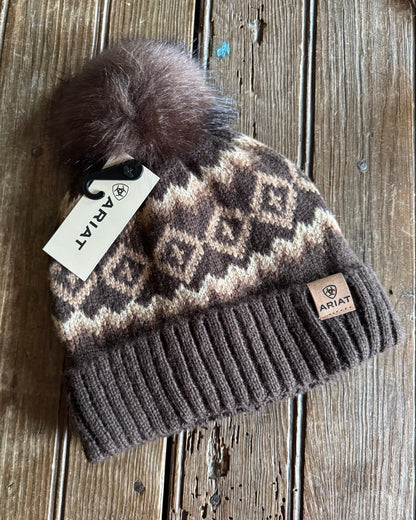 Brown beanie with patterned design and pom-pom on a wooden surface, featuring Ariat branding.
