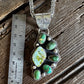 A silver-toned paw print pendant necklace with green and turquoise stones, displayed on a rustic wooden surface next to a metal ruler for scale.