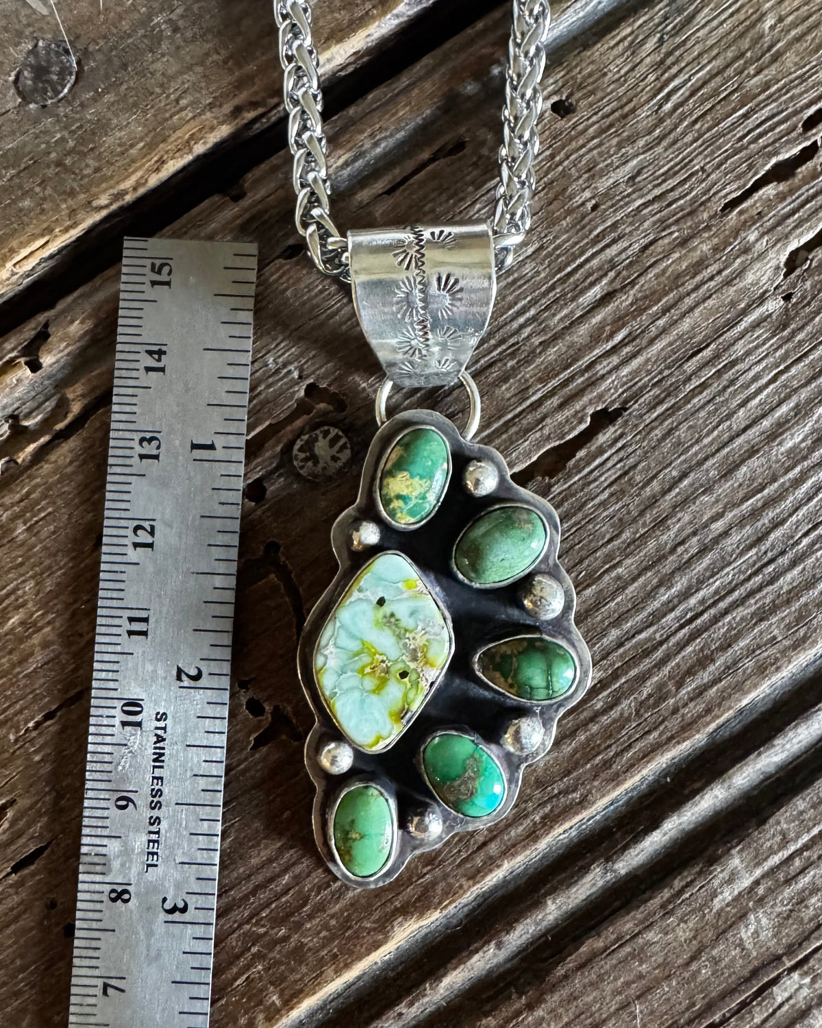 A silver-toned paw print pendant necklace with green and turquoise stones, displayed on a rustic wooden surface next to a metal ruler for scale.