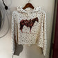 A cream-colored cropped hoodie with a small floral print and a large brown horse graphic on the front, hanging on a wooden hanger against a white wall.