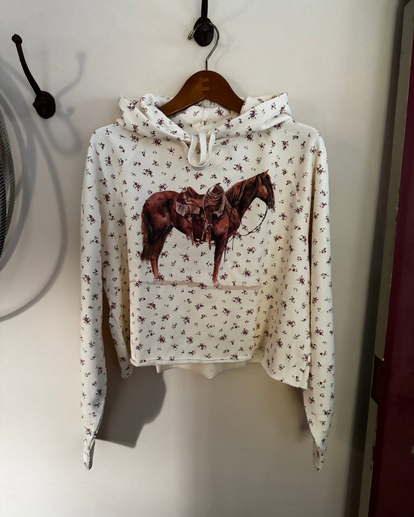 A cream-colored cropped hoodie with a small floral print and a large brown horse graphic on the front, hanging on a wooden hanger against a white wall.