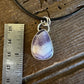 A silver pendant necklace with a teardrop-shaped purple and lavender agate stone, displayed on a rustic wooden surface next to a metal ruler for scale.