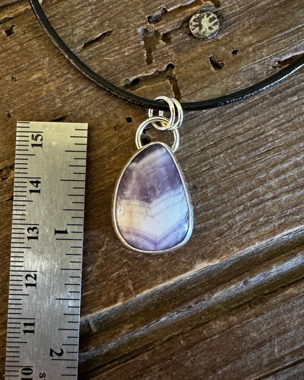 A silver pendant necklace with a teardrop-shaped purple and lavender agate stone, displayed on a rustic wooden surface next to a metal ruler for scale.