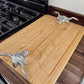 A rectangular bamboo cutting board with a carved handle slot, featuring two decorative silver metal handles shaped like longhorn cattle, shown on a kitchen counter next to a black gas stove.