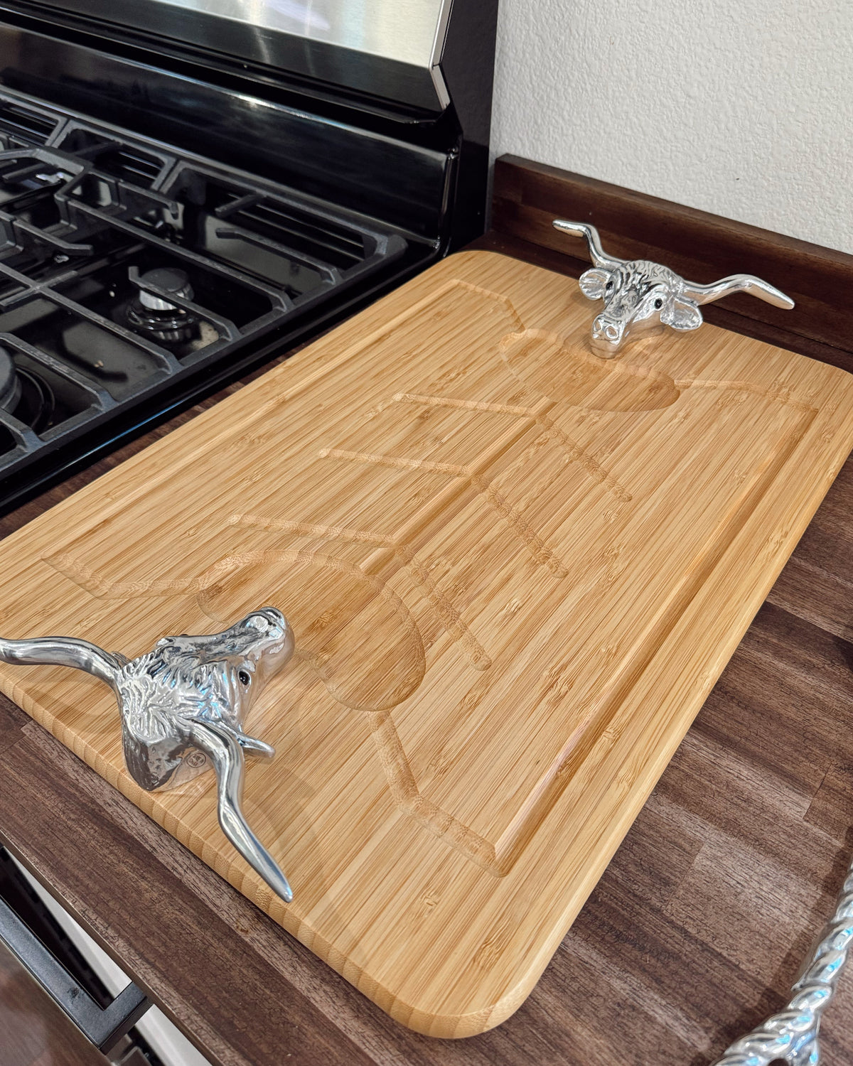 A rectangular bamboo cutting board with a carved handle slot, featuring two decorative silver metal handles shaped like longhorn cattle, shown on a kitchen counter next to a black gas stove.