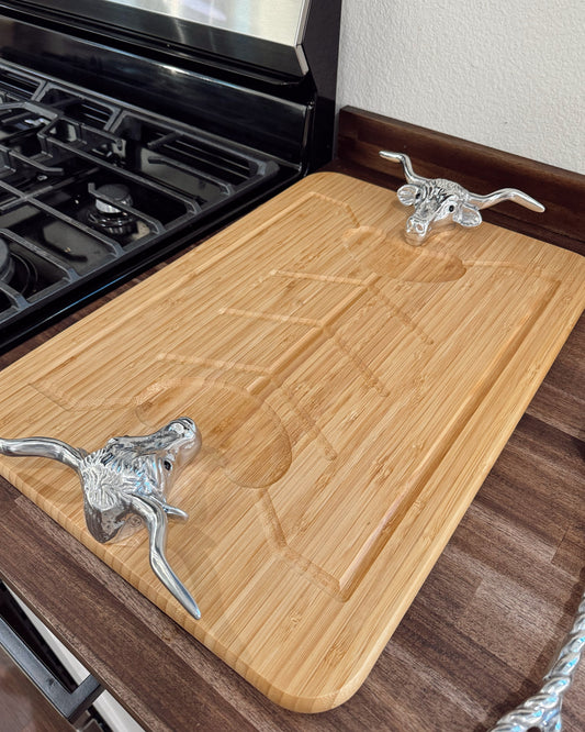 A rectangular bamboo cutting board with a carved handle slot, featuring two decorative silver metal handles shaped like longhorn cattle, shown on a kitchen counter next to a black gas stove.