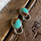 A pair of silver-toned drop earrings with oval turquoise and brown stone inlays, displayed on a rustic wooden surface next to a white faux fur fabric.
