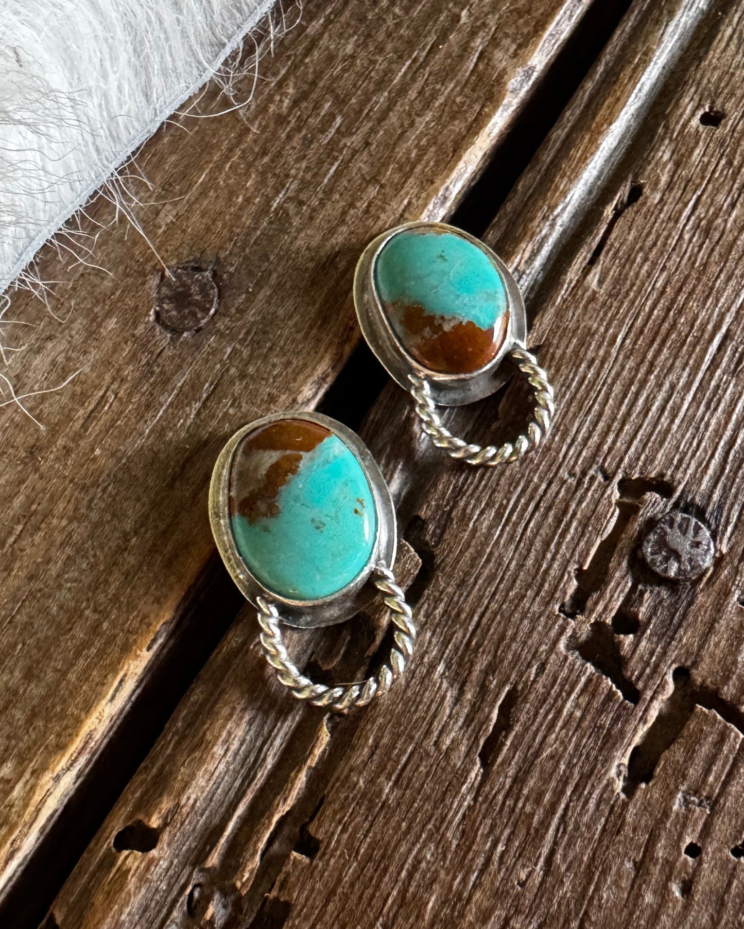 A pair of silver-toned drop earrings with oval turquoise and brown stone inlays, displayed on a rustic wooden surface next to a white faux fur fabric.