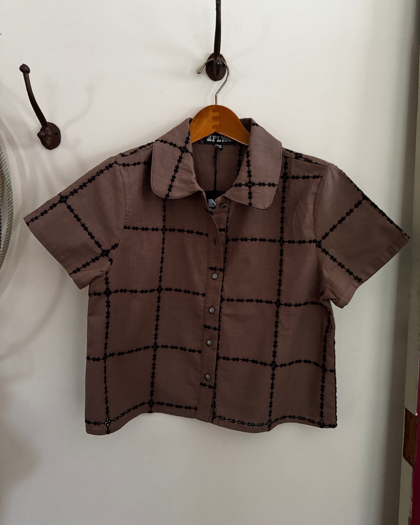 A short-sleeve button-up shirt in brown with a black grid pattern, hanging on a wooden hanger against a white wall.