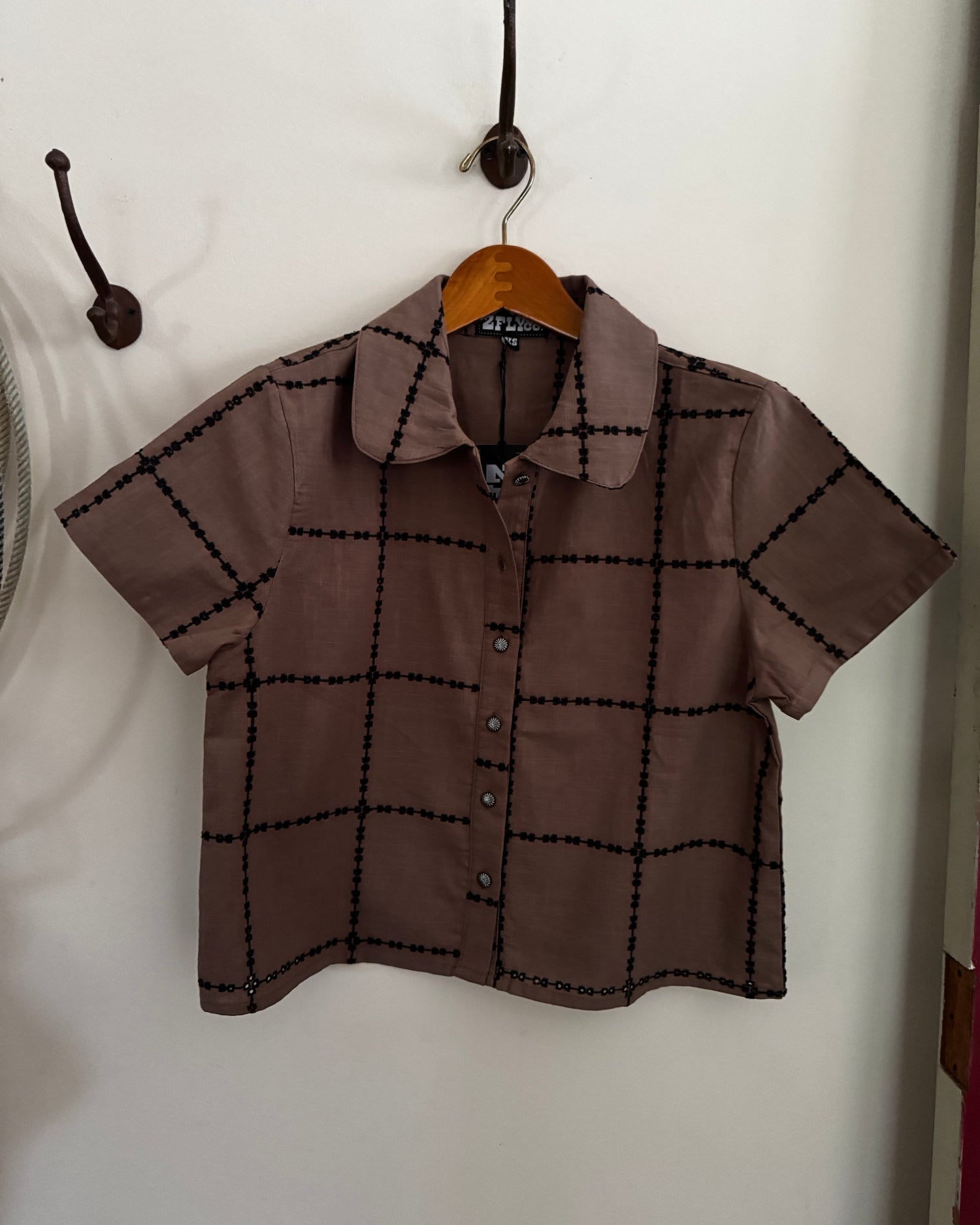A short-sleeve button-up shirt in brown with a black grid pattern, hanging on a wooden hanger against a white wall.