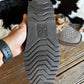 A hand holds up the treaded sole of a dark brown boot or shoe, showing the ARIAT brand logo embossed in the center. The background features a rustic wooden floor and a cowhide rug.