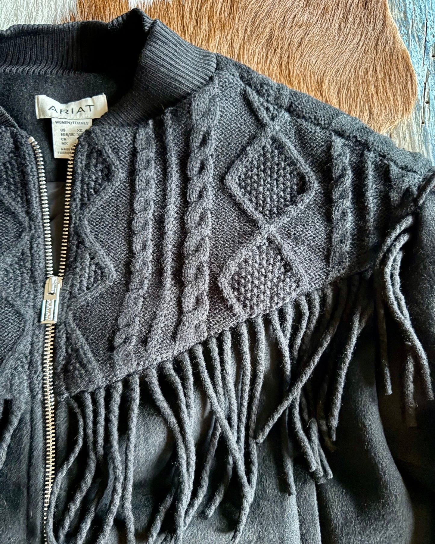 Black zip-up bomber jacket with intricate pattern and fringes on a wooden surface