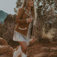 Woman in a brown jacket and white boots standing on rocky terrain with mountains in the background