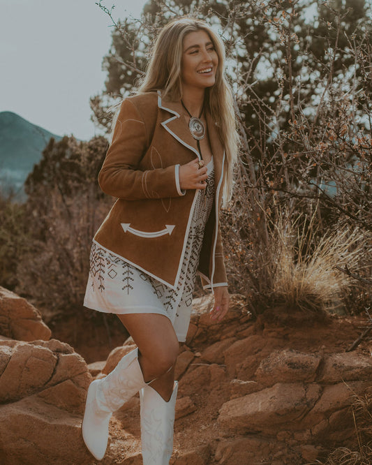 Woman in a brown jacket and white boots standing on rocky terrain with mountains in the background