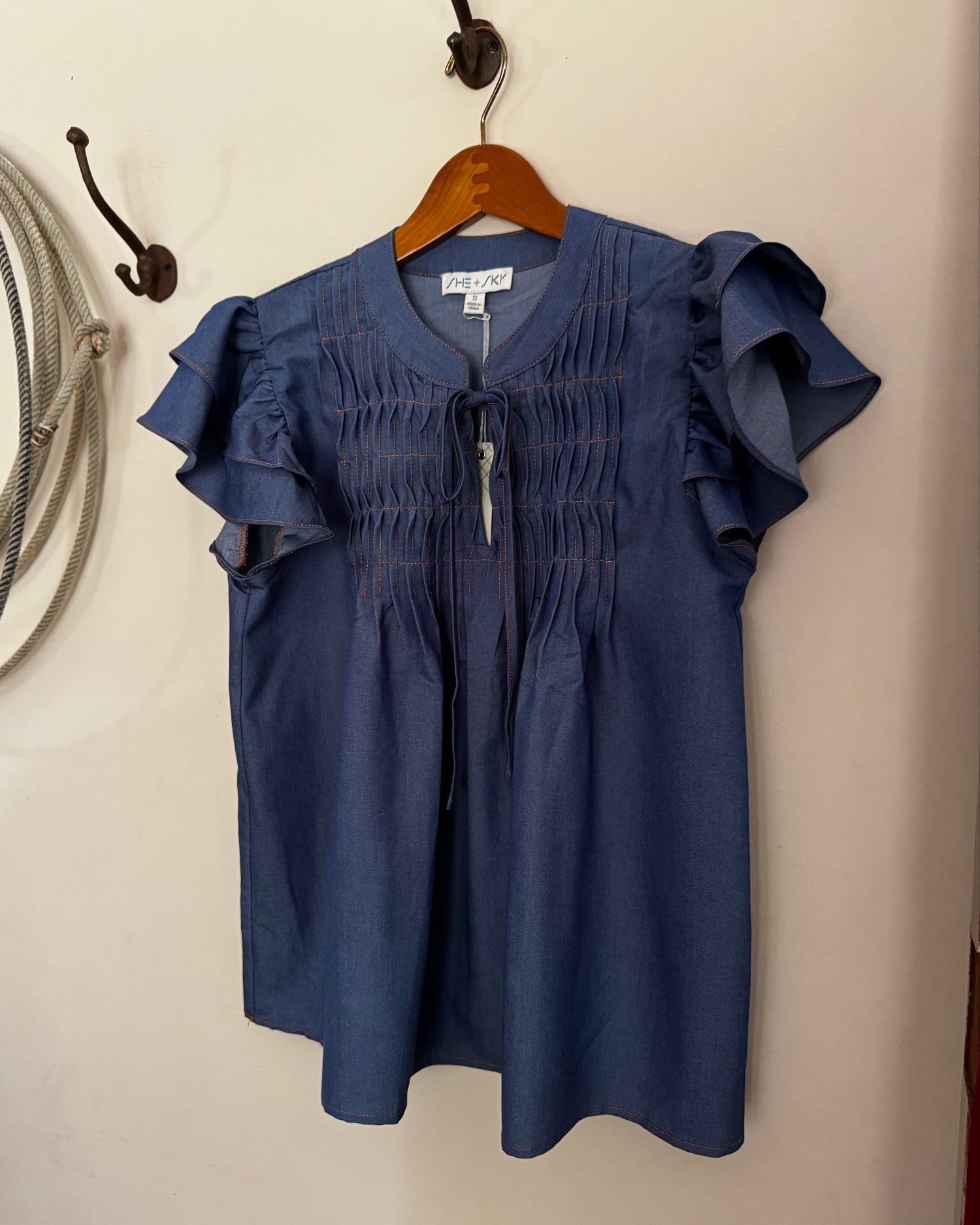 Blue blouse with ruffled sleeves on a hanger against a beige wall.