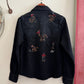 Black denim shirt with embroidered designs of horses and people on a white wall.