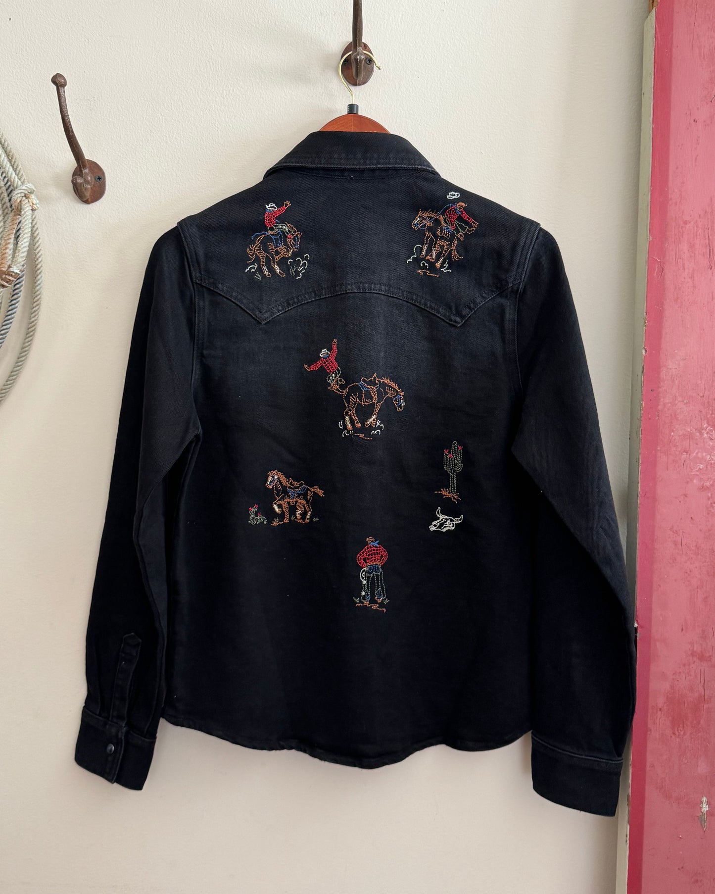 Black denim shirt with embroidered designs of horses and people on a white wall.
