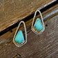 A pair of teardrop-shaped turquoise earrings with textured silver borders, displayed on a rustic dark wood surface with a white faux fur accent at the top.