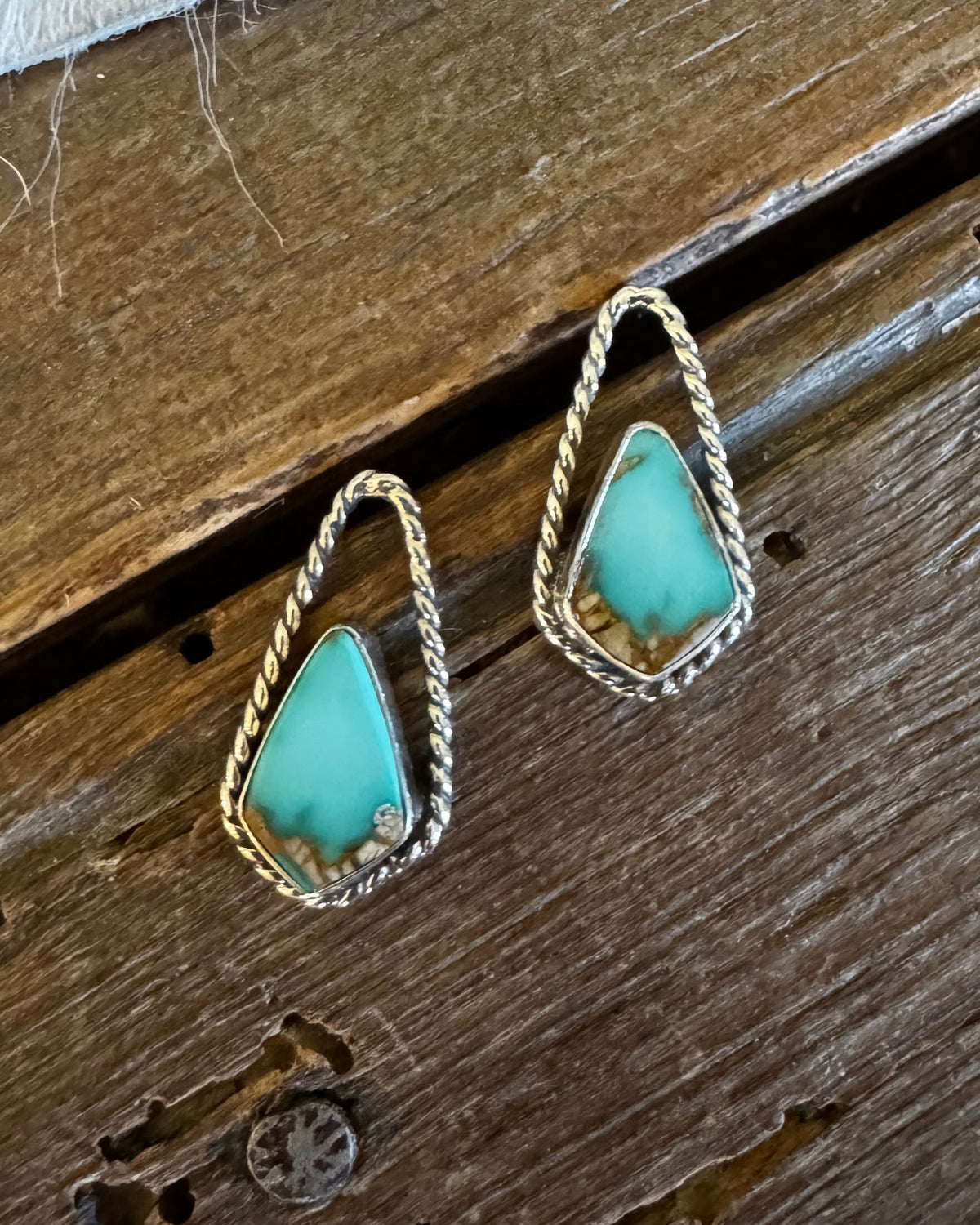 A pair of teardrop-shaped turquoise earrings with textured silver borders, displayed on a rustic dark wood surface with a white faux fur accent at the top.