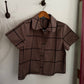 A short-sleeve collared button-up shirt in a muted taupe-brown color with a black windowpane plaid pattern, hanging on a wooden hanger from a wall hook.