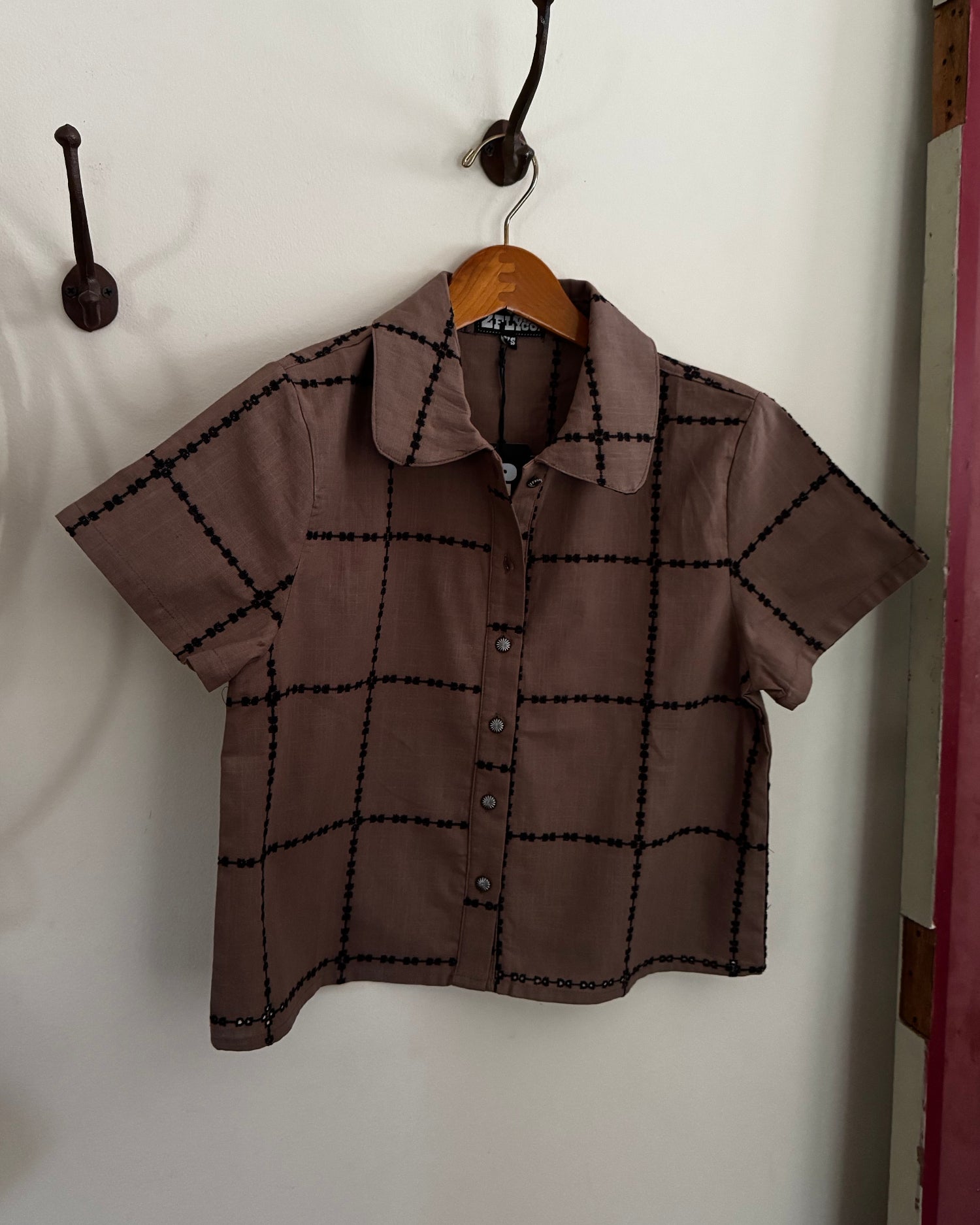 A short-sleeve collared button-up shirt in a muted taupe-brown color with a black windowpane plaid pattern, hanging on a wooden hanger from a wall hook.