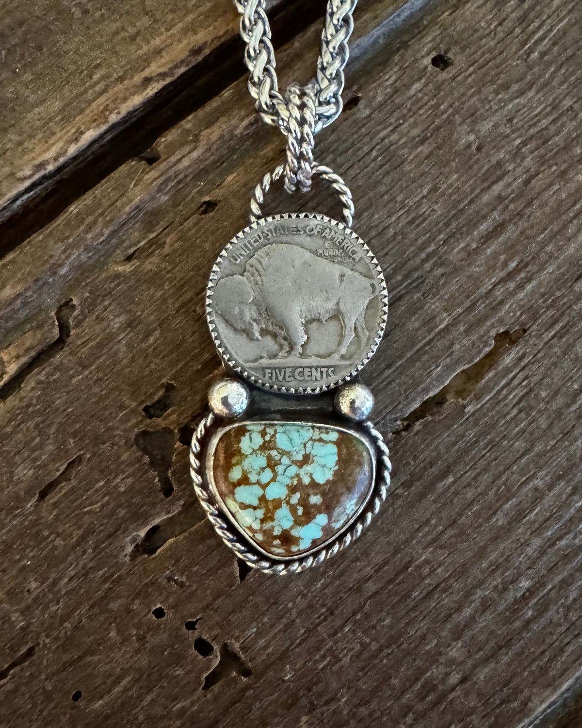 A silver-toned necklace featuring a circular coin pendant and a heart-shaped turquoise stone pendant, displayed on a rustic dark wood surface.