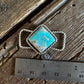 A vintage-style silver twilly slide with a square turquoise cabochon and twisted rope band, shown next to a stainless steel ruler on a rustic wooden surface.