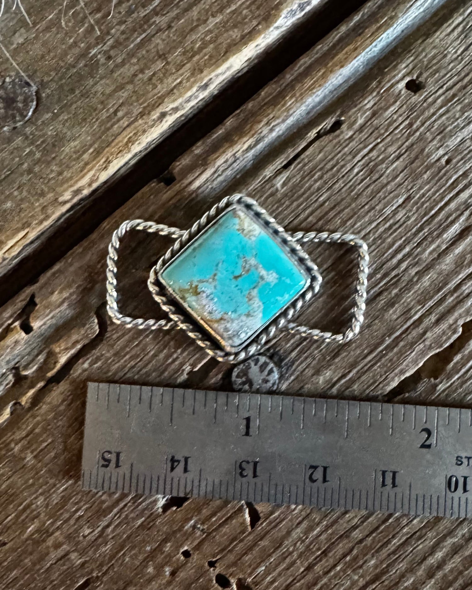 A vintage-style silver twilly slide with a square turquoise cabochon and twisted rope band, shown next to a stainless steel ruler on a rustic wooden surface.