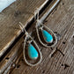 A pair of silver-toned teardrop dangle earrings with vibrant turquoise stone centers, displayed on a rustic dark wood surface.