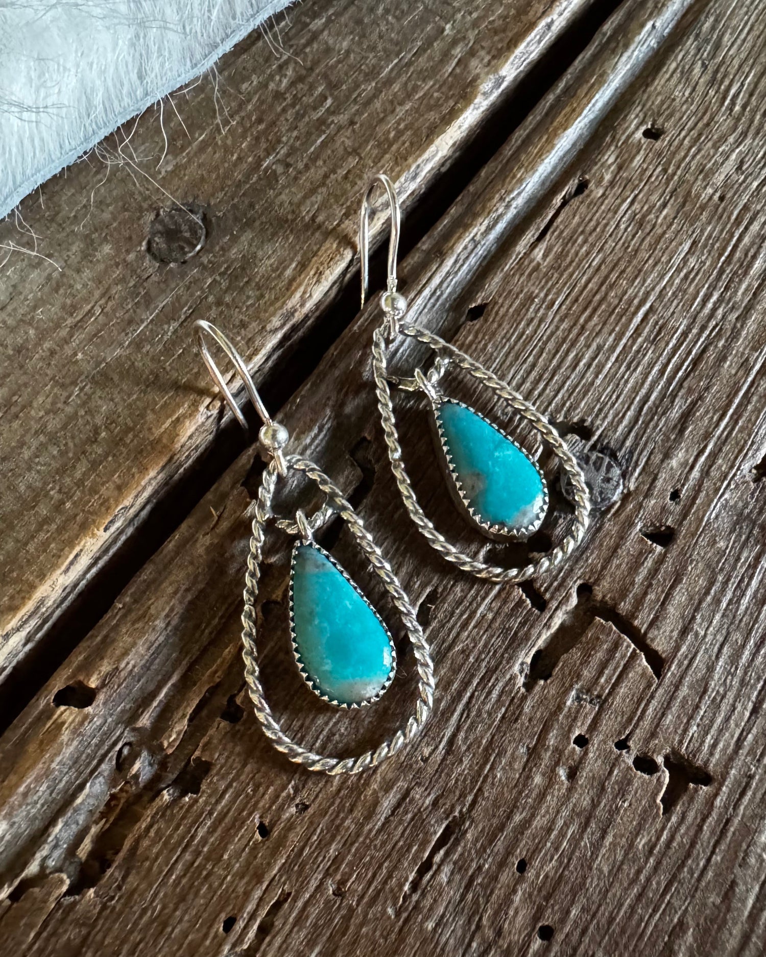 A pair of silver-toned teardrop dangle earrings with vibrant turquoise stone centers, displayed on a rustic dark wood surface.
