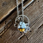 A silver-toned necklace with a unique teardrop pendant featuring a blue and white marbled top half and a golden-yellow patterned bottom half, displayed on a rustic dark wood background.