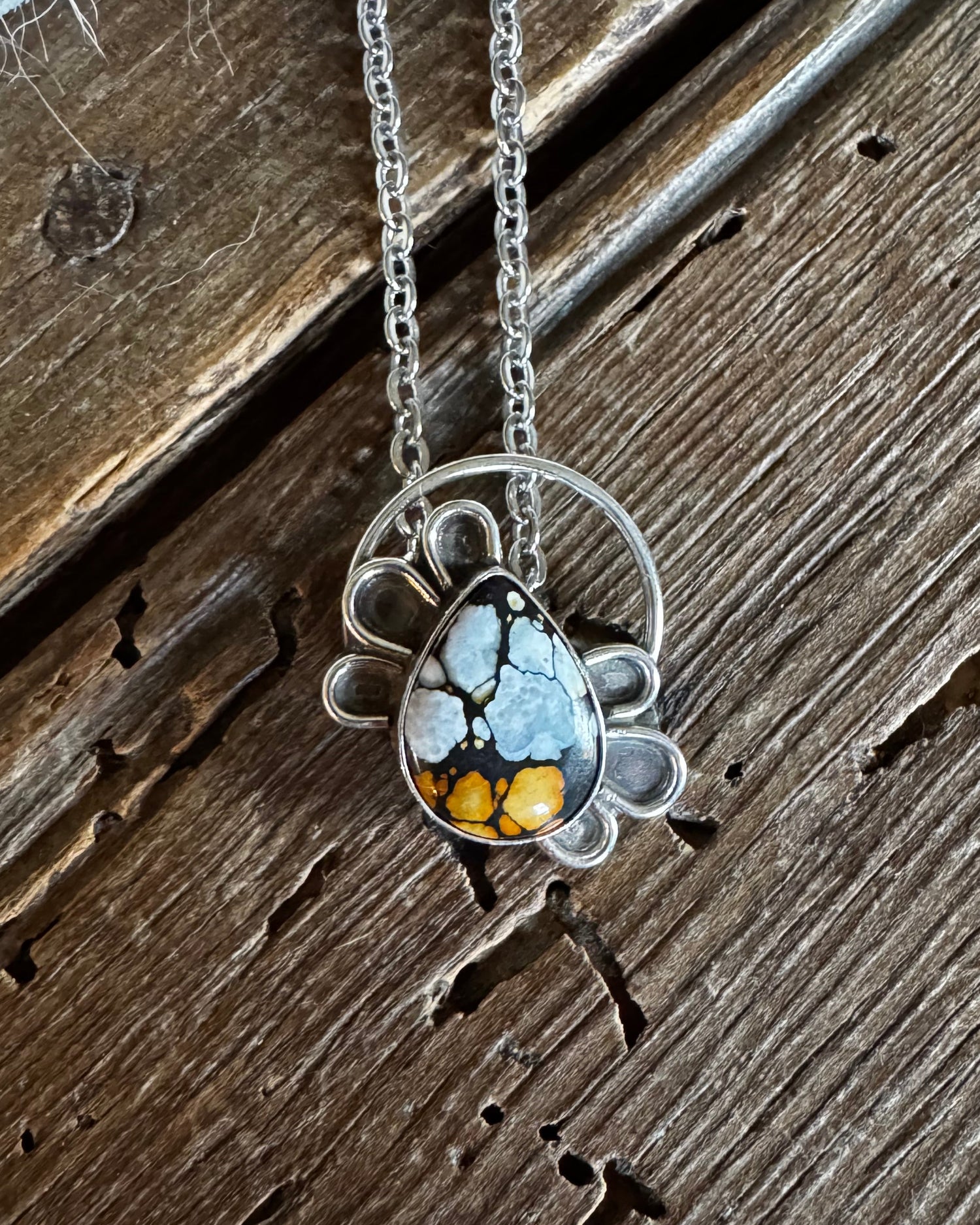 A silver-toned necklace with a unique teardrop pendant featuring a blue and white marbled top half and a golden-yellow patterned bottom half, displayed on a rustic dark wood background.
