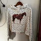 A white cropped hoodie with a brown horse graphic on the front, featuring a small all-over floral print, hanging on a wooden hanger against a beige wall.