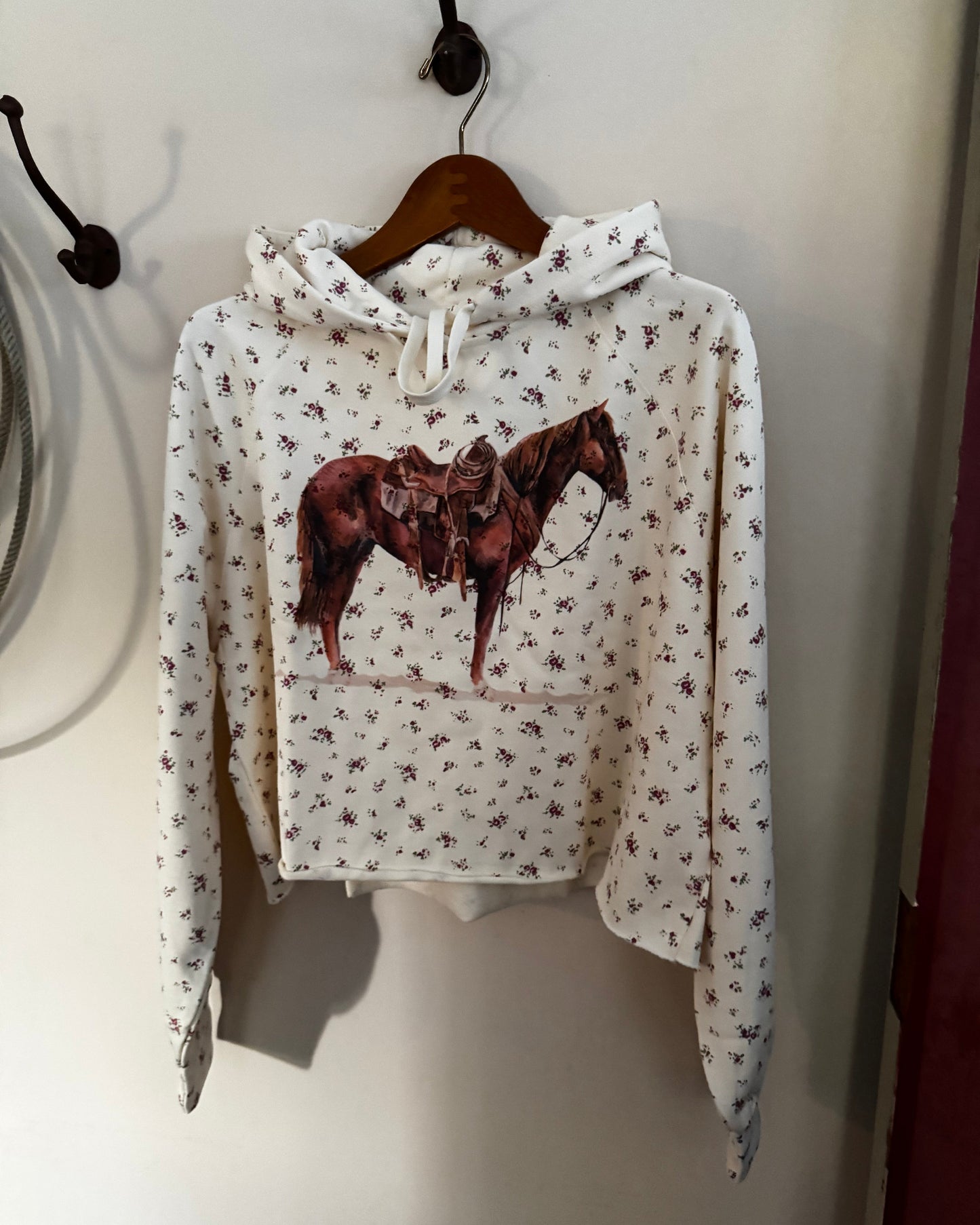 A white cropped hoodie with a brown horse graphic on the front, featuring a small all-over floral print, hanging on a wooden hanger against a beige wall.