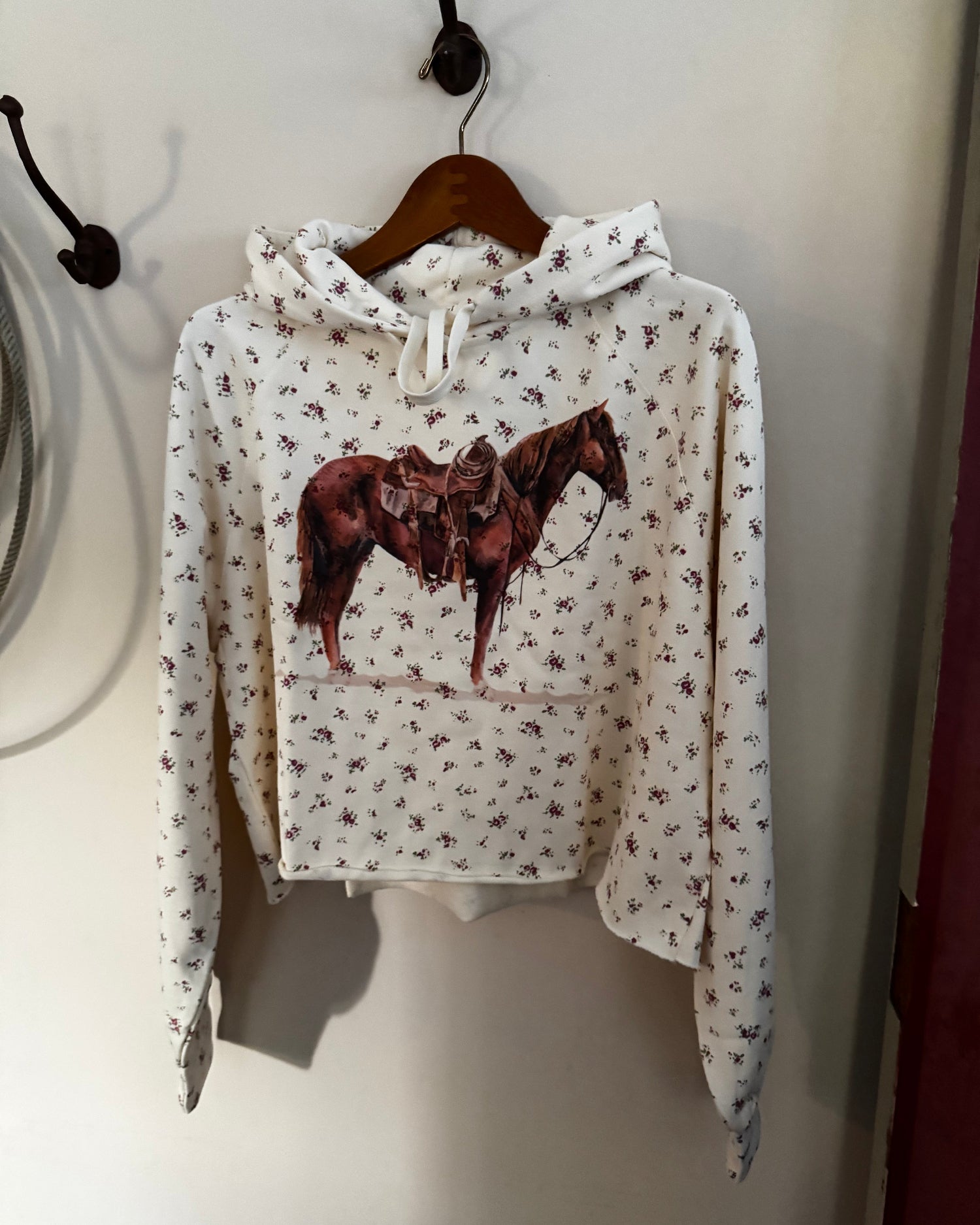 A white cropped hoodie with a brown horse graphic on the front, featuring a small all-over floral print, hanging on a wooden hanger against a beige wall.