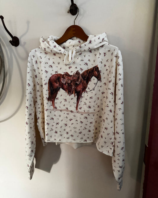 A white cropped hoodie with a brown horse graphic on the front, featuring a small all-over floral print, hanging on a wooden hanger against a beige wall.