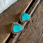A pair of triangular turquoise stud earrings with silver-toned bezel settings, resting on rustic wooden planks next to white fur.