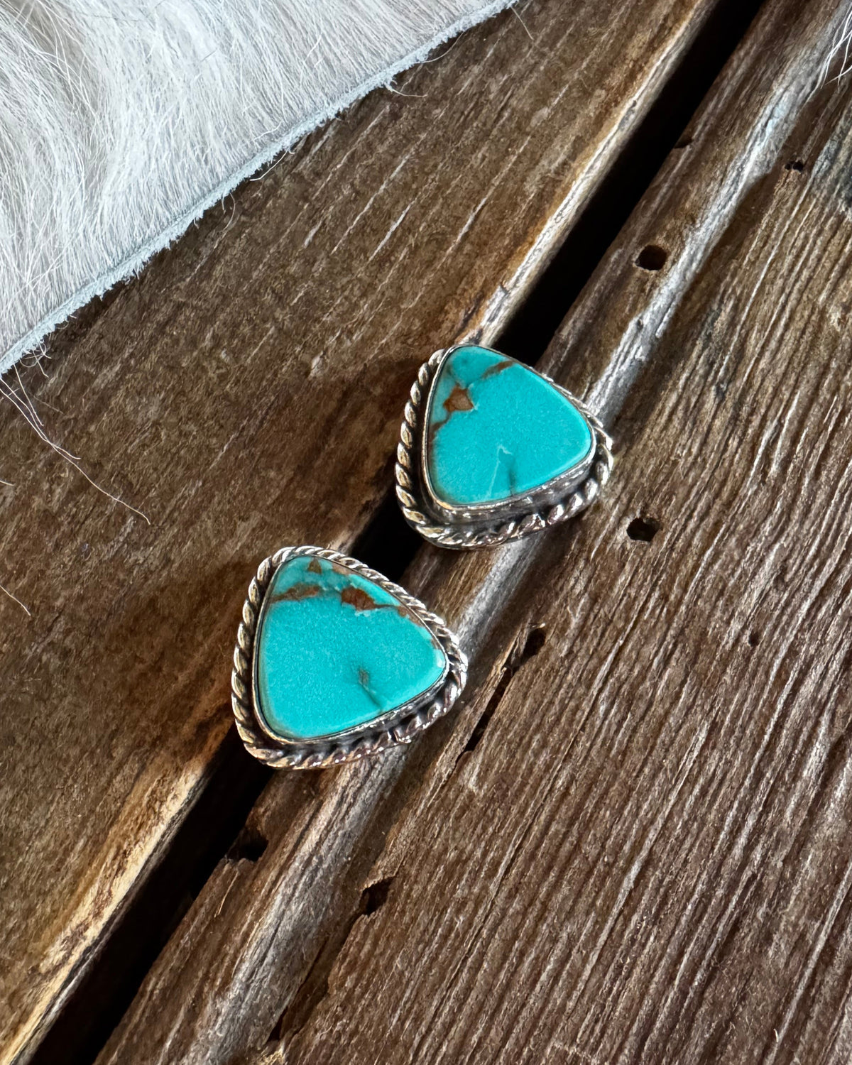 A pair of triangular turquoise stud earrings with silver-toned bezel settings, resting on rustic wooden planks next to white fur.