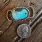 Turquoise stone pendant with silver frame on a wooden surface next to a penny for scale.