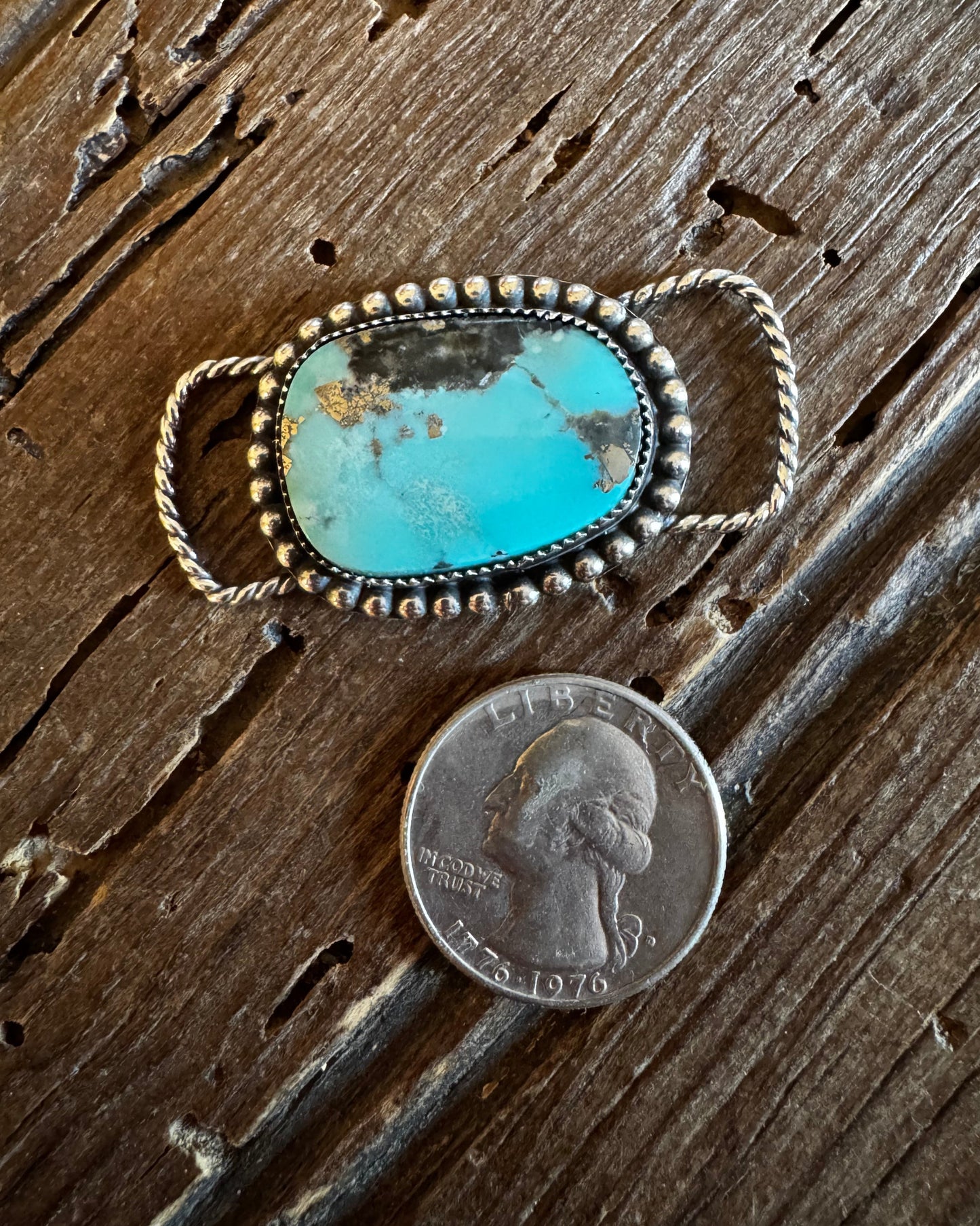 Turquoise stone pendant with silver frame on a wooden surface next to a penny for scale.