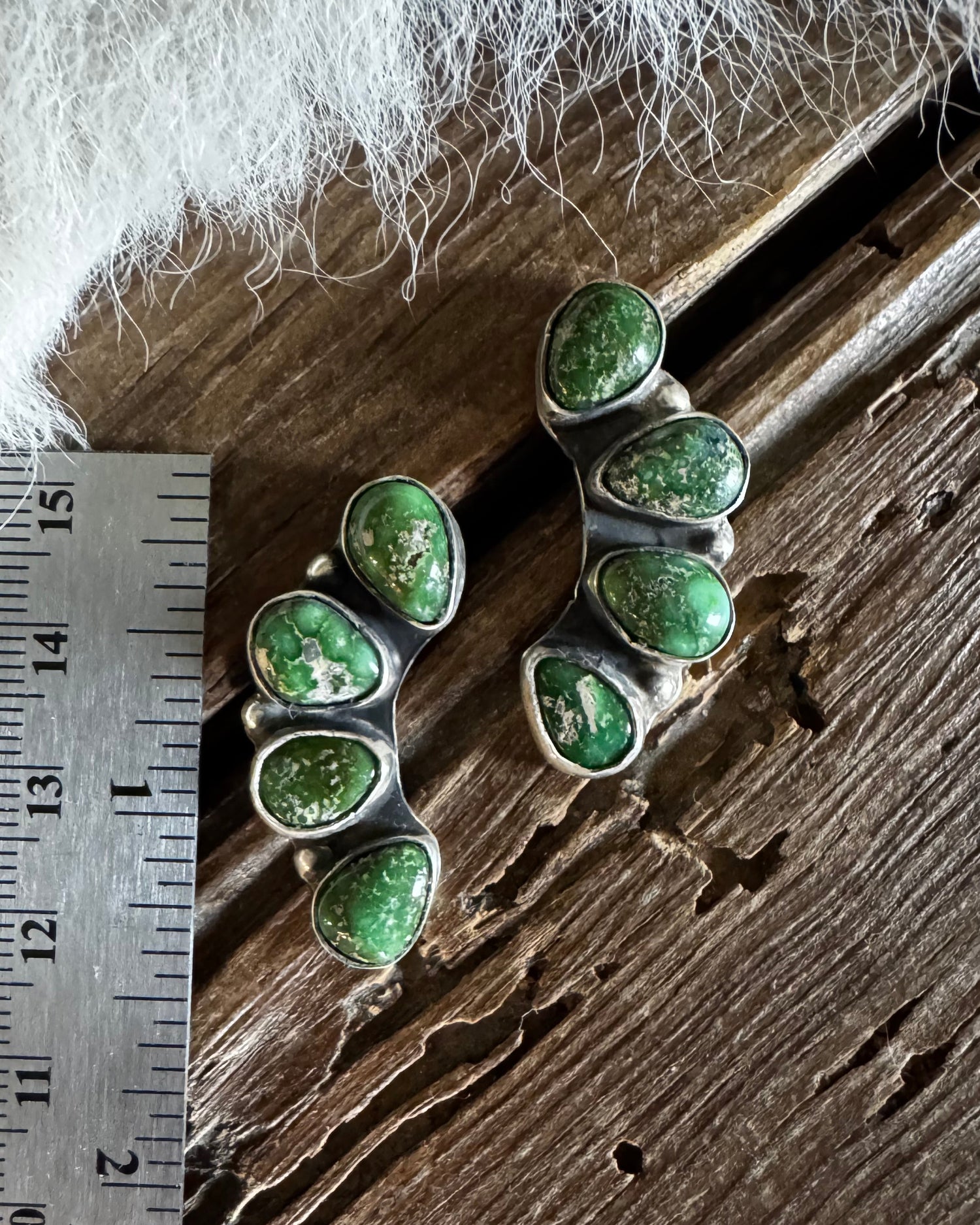 Two silver earrings with green gemstones on a wooden surface with a ruler for scale.