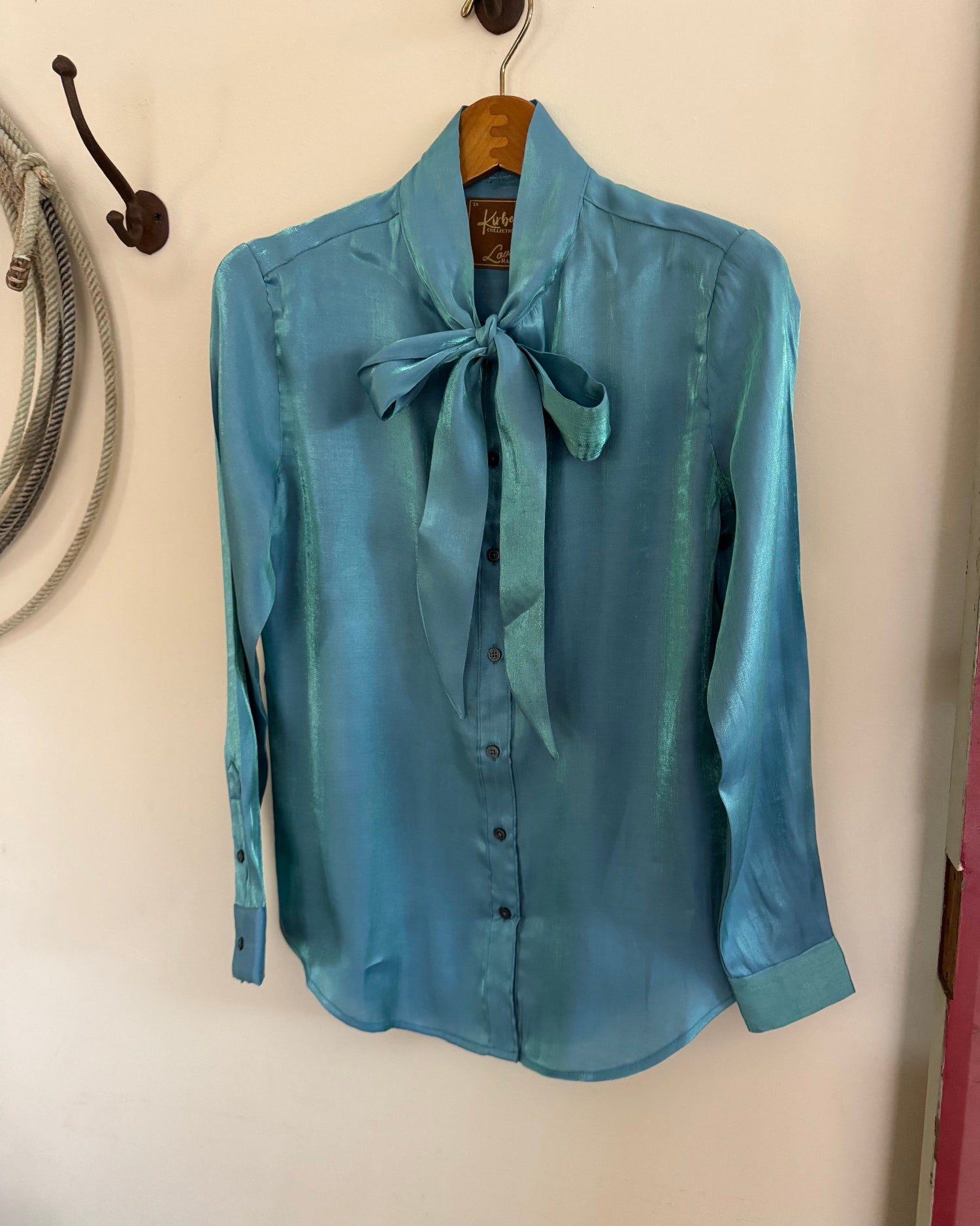 Blue blouse with a tie collar hanging on a hook against a white wall.