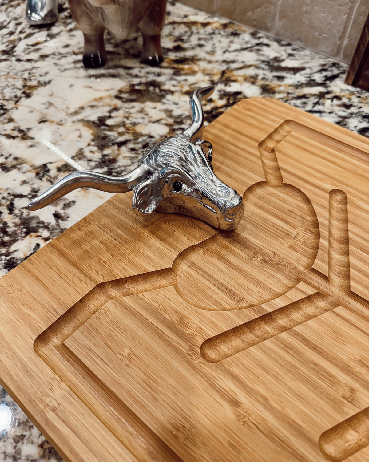 A silver bull head cheese knife resting on a rectangular bamboo cutting board with a geometric design, set on a speckled granite countertop.
