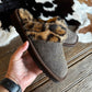 A person's hand holds a brown felt slipper with a leopard-print faux fur trim and small silver studs, set against a rustic wood floor and a cowhide rug.
