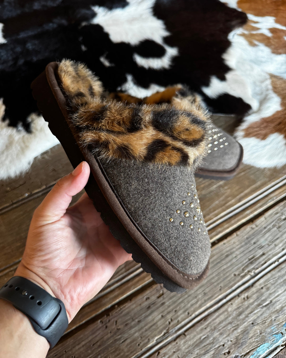 A person's hand holds a brown felt slipper with a leopard-print faux fur trim and small silver studs, set against a rustic wood floor and a cowhide rug.