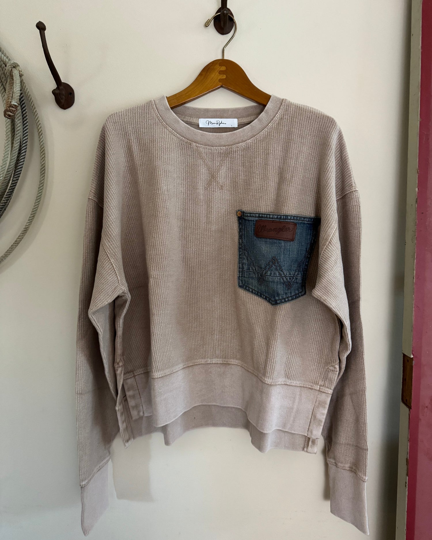 Beige sweatshirt with a denim pocket on a wooden hanger against a white wall.