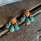 A pair of silver stud earrings with turquoise and orange stone accents, displayed on a rustic wooden surface with white faux fur in the background.