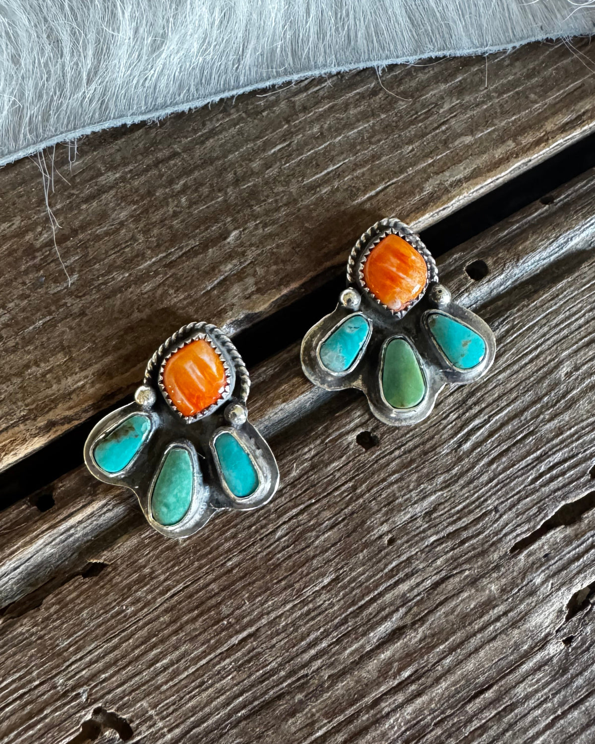 A pair of silver stud earrings with turquoise and orange stone accents, displayed on a rustic wooden surface with white faux fur in the background.