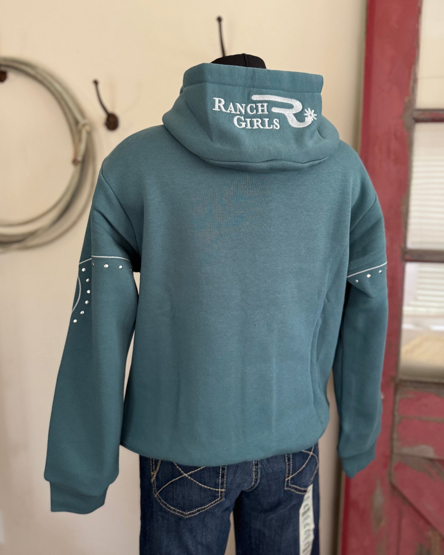 Blue hoodie with 'Ranch Girls' logo on a person standing against a wall with a red door and coiled rope.