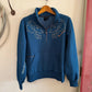 Blue hoodie with floral embroidery on a wooden hanger against a white wall.